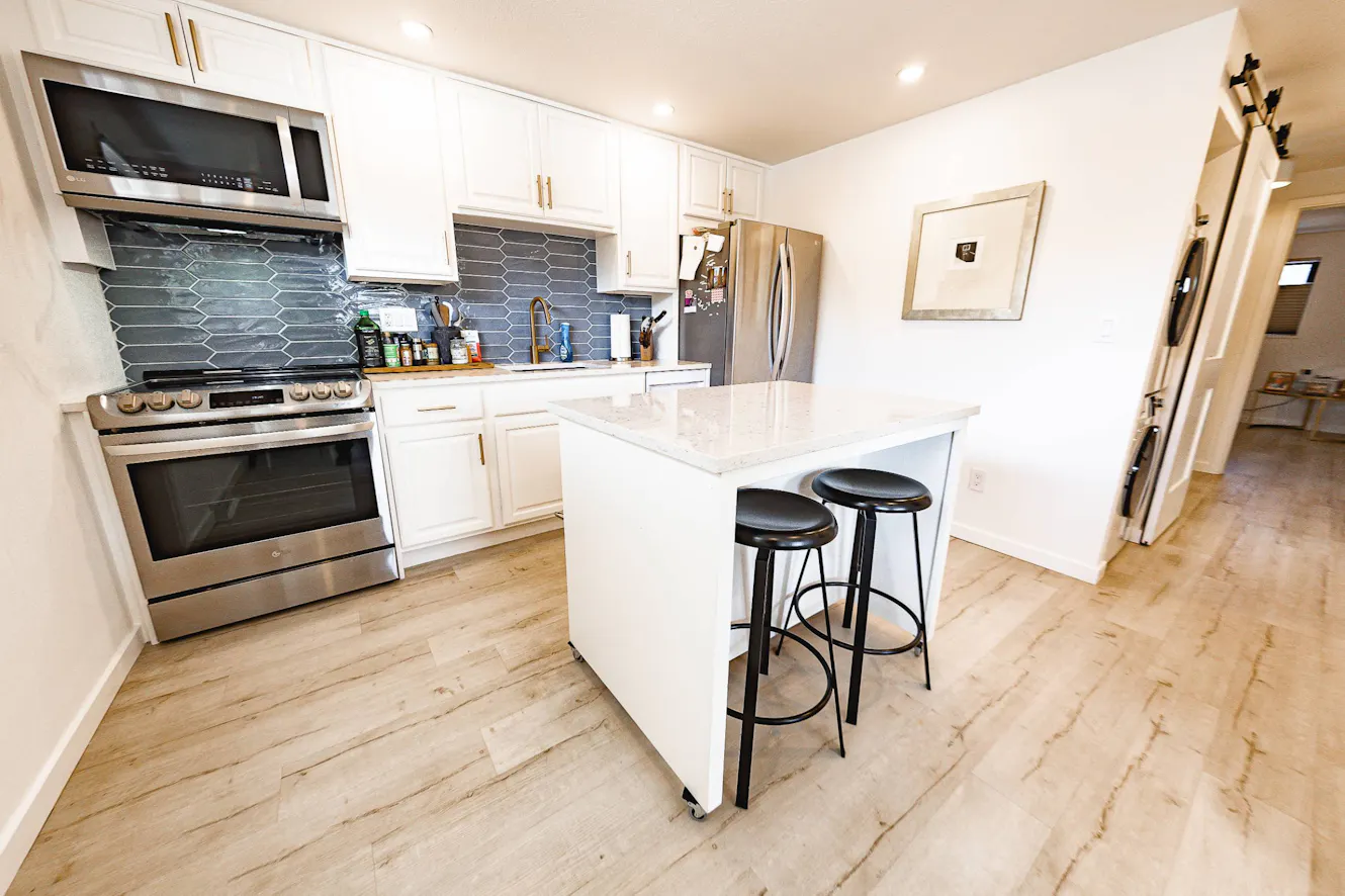 Kitchen with white cabinets, stainless steel appliances, gray hexagonal tile backsplash, white quartz countertops, and breakfast bar with two black stools