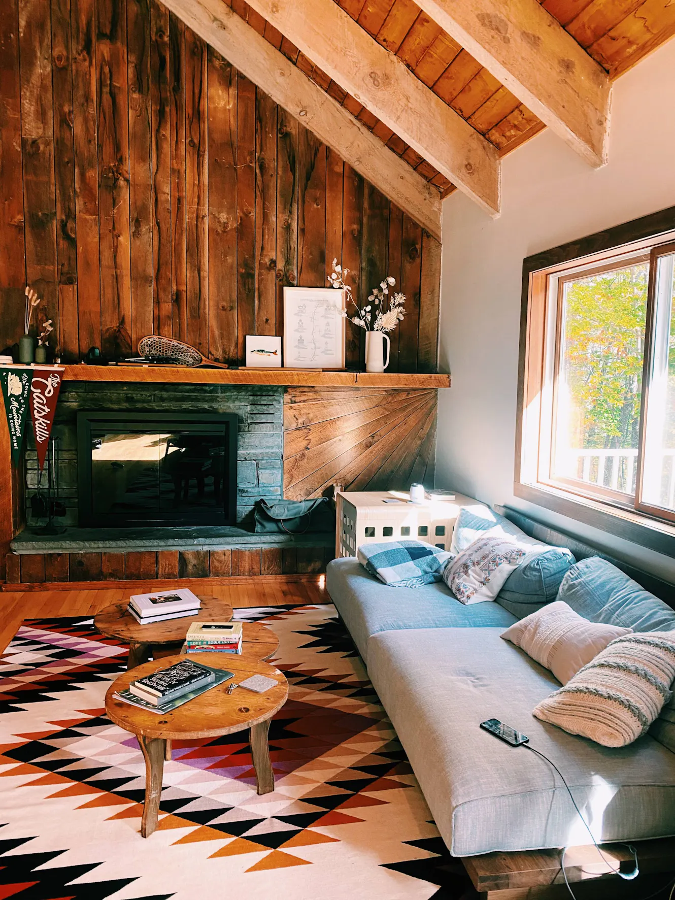 Living room with vaulted wood ceiling, stone fireplace with wooden mantel, gray sectional sofa, round coffee table, geometric patterned rug, and large window with tree views