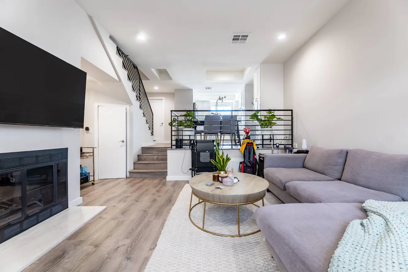 Living room with gray sectional sofa, round coffee table, wall-mounted TV, fireplace, staircase with metal railing, and workspace area with plants