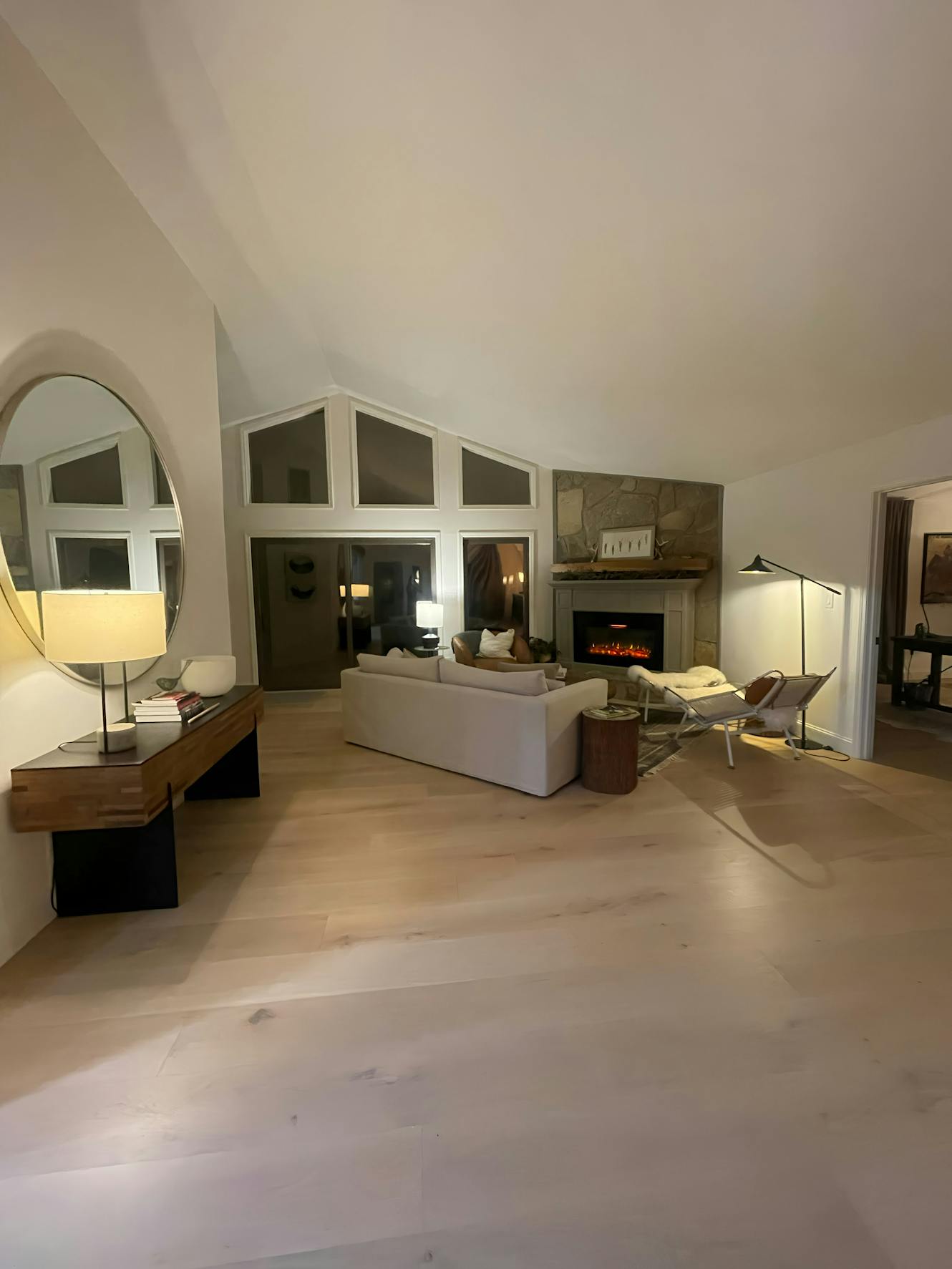 Living room with vaulted ceiling, white sectional sofa, stone fireplace, light wood floors, arched mirror, and modern lounge chair