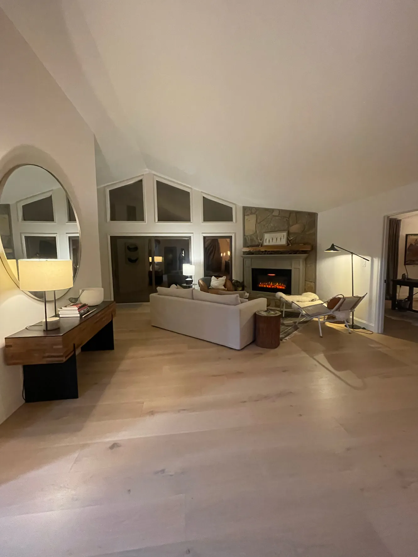 Living room with vaulted ceiling, white sectional sofa, stone fireplace, light wood floors, arched mirror, and modern lounge chair