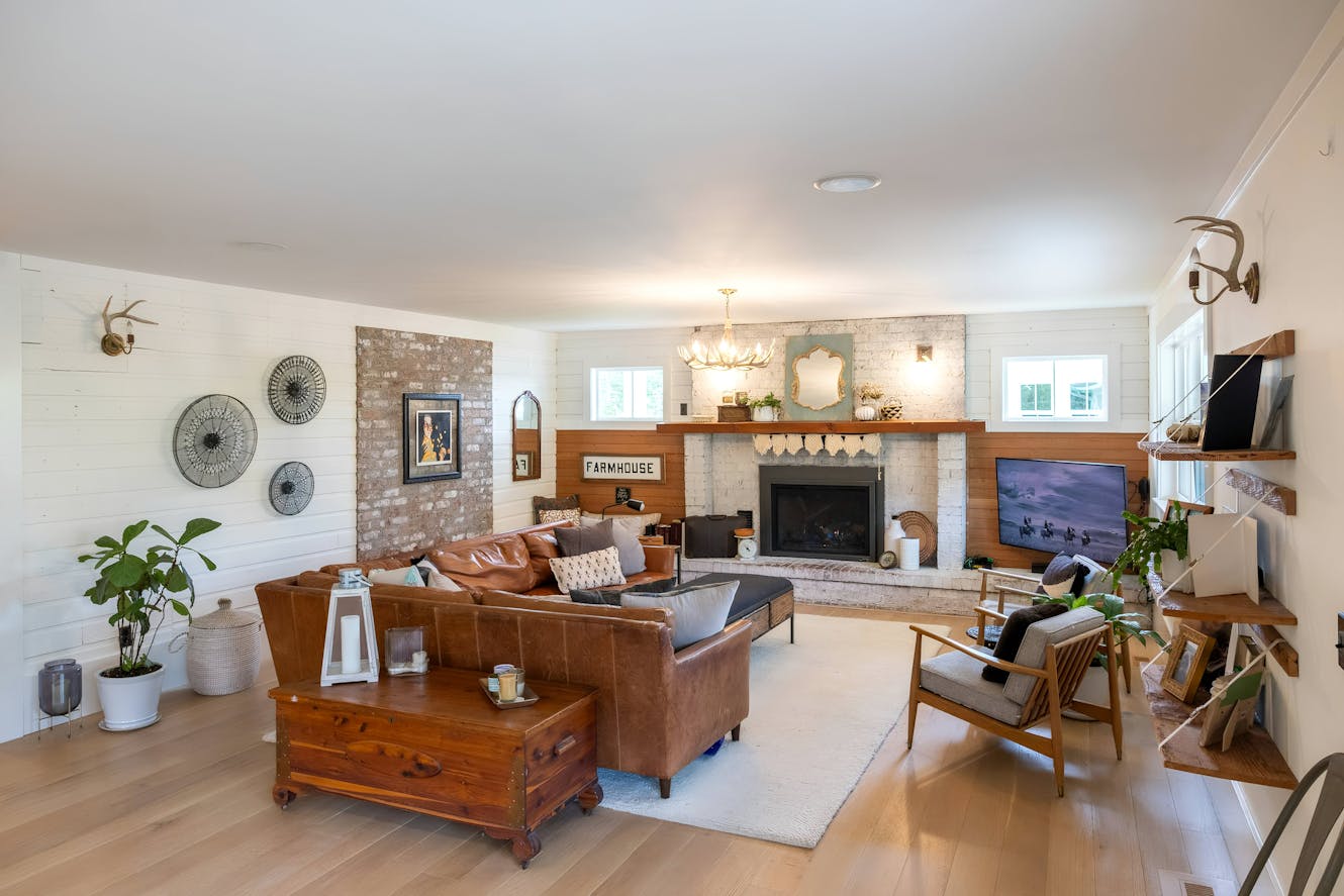 Living room with brown leather sofas, exposed brick fireplace with wood mantel, white shiplap walls, wooden coffee table trunk, and wall-mounted TV