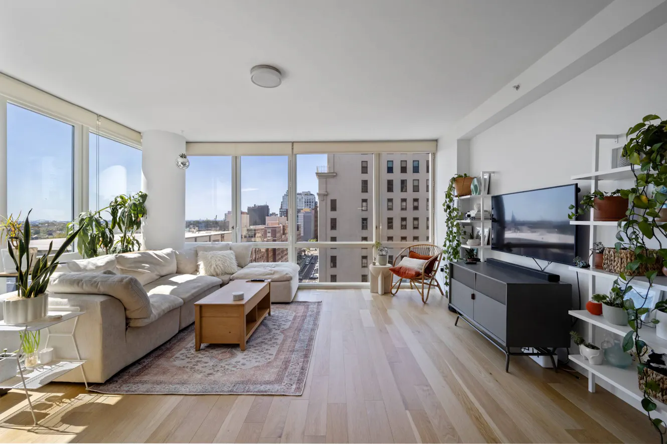 Living room with beige sectional sofa, wooden coffee table, large floor-to-ceiling windows with city views, TV on media console, and multiple potted plants