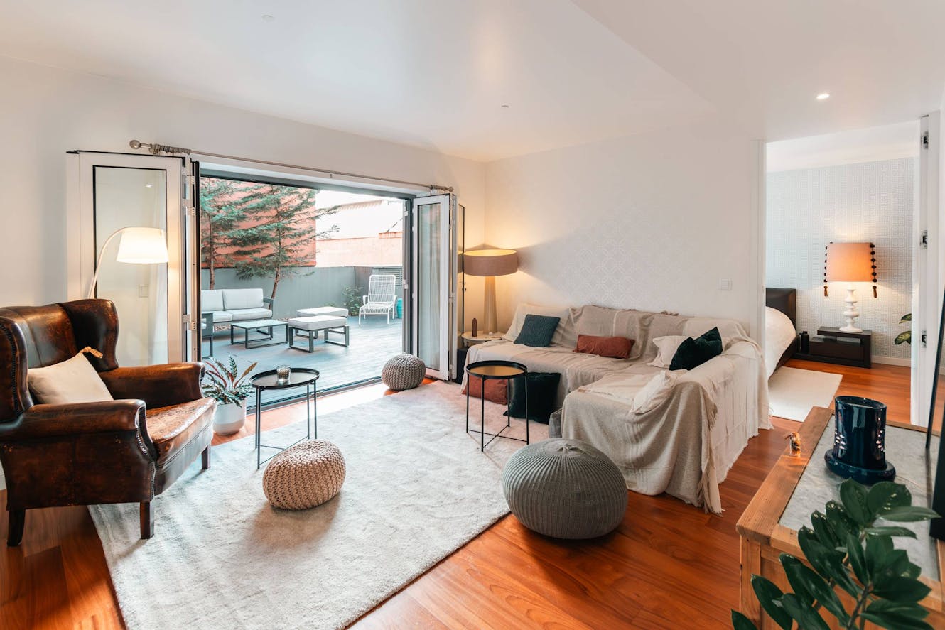 Living room with beige sectional sofa, brown leather armchair, hardwood floors, area rug, and folding glass doors opening to patio with outdoor seating