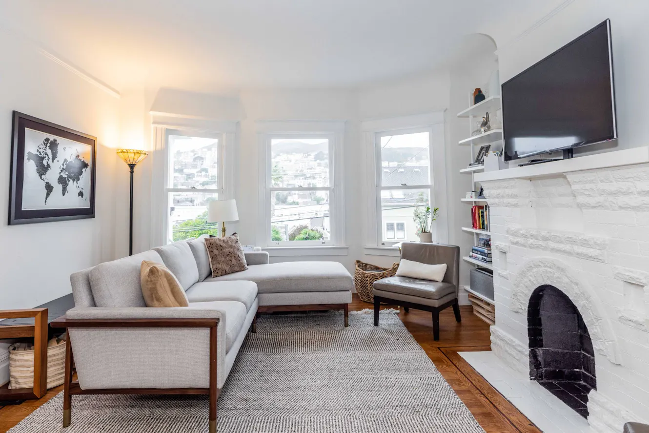 Living room with gray sectional sofa, three large windows with natural light, white brick fireplace, wall-mounted TV, and hardwood floors