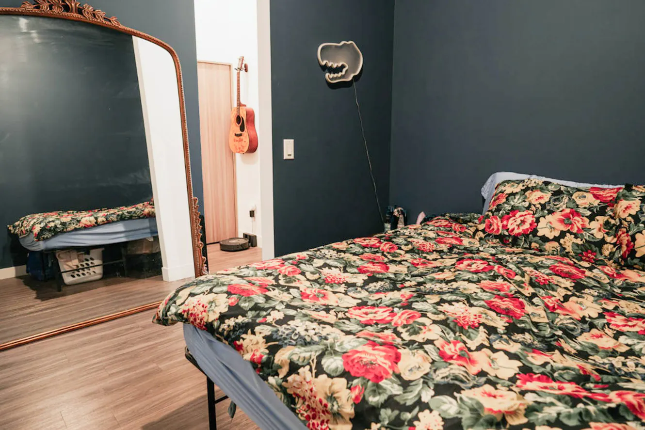 Bedroom with queen bed with floral bedding, dark teal walls, large ornate mirror, and neon dinosaur wall light