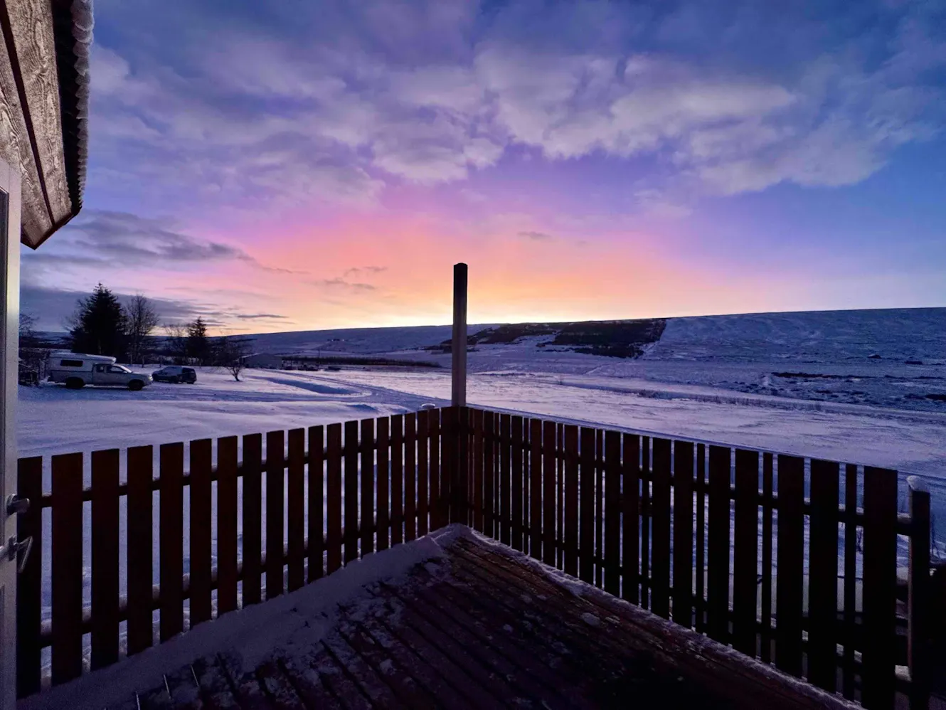 Patio or balcony with wooden fence overlooking snow-covered landscape at sunset with pink and blue sky