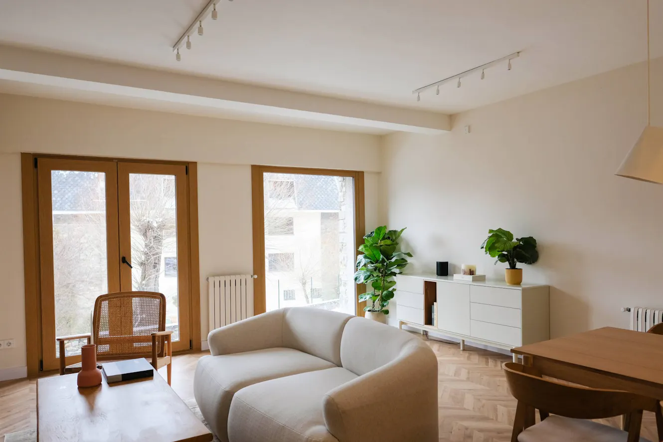 Living room with beige curved sofa, wooden French doors, white credenza with plants, cane accent chair, and herringbone wood flooring