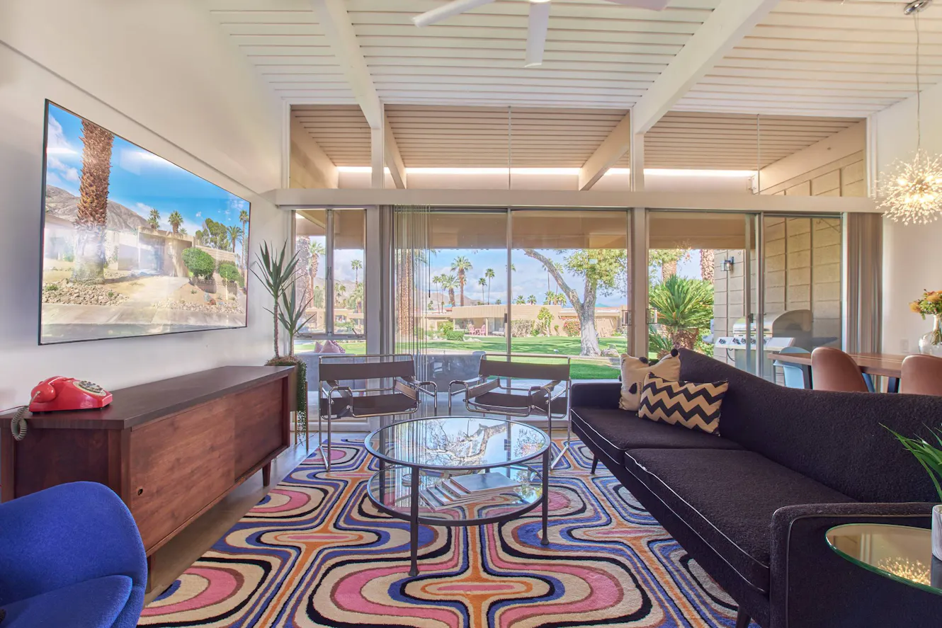 Living room with dark gray sofa, glass coffee table, mid-century modern chairs, colorful patterned rug, wall-mounted TV, and floor-to-ceiling windows overlooking backyard with palm trees