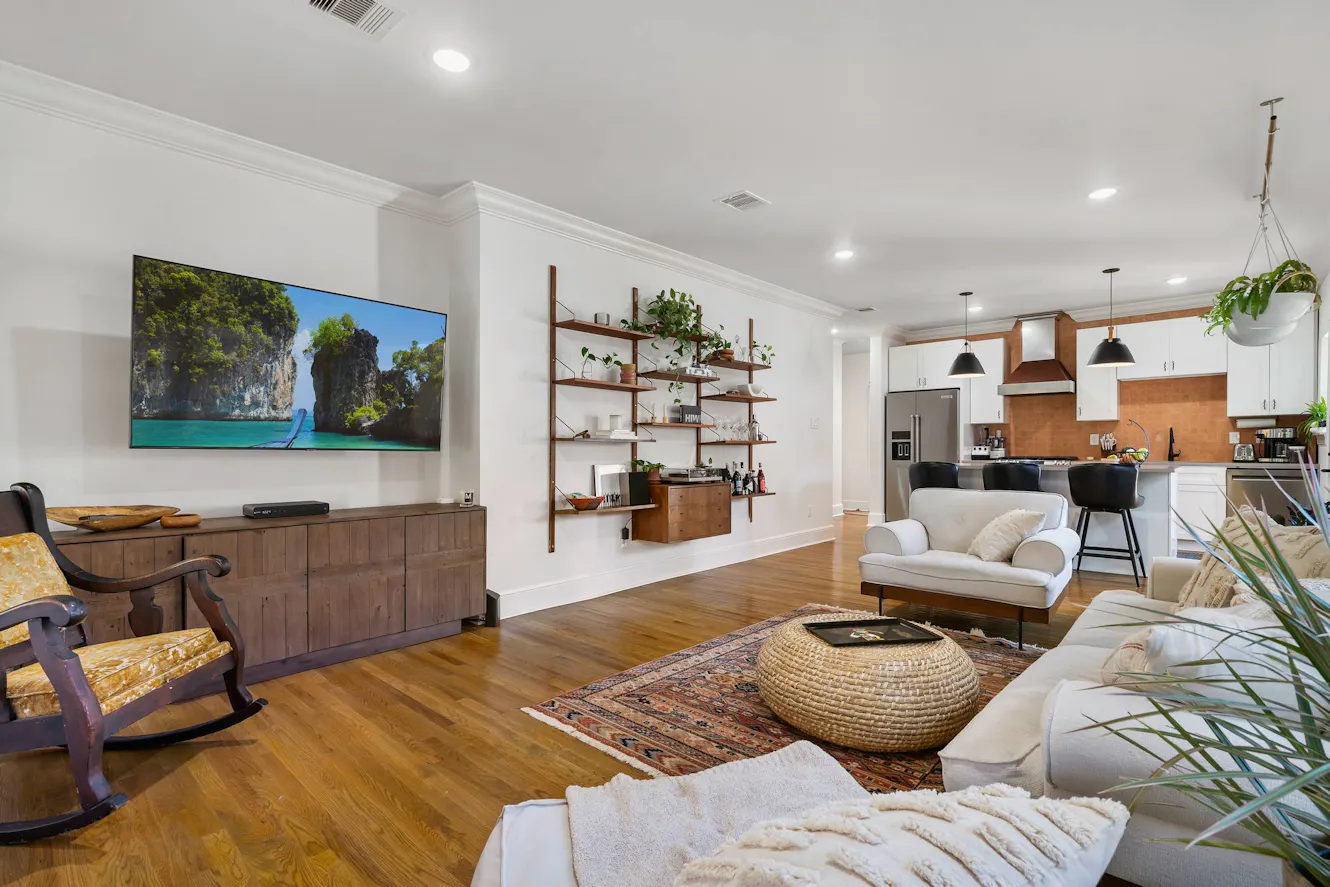 Open-concept living room and kitchen with white sectional sofa, wall-mounted TV, wooden media console, wall shelving with plants, hardwood floors, and kitchen with white cabinets