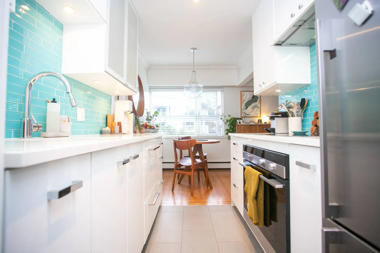 Kitchen with white cabinets, turquoise subway tile backsplash, stainless steel appliances, white countertops, and small dining table with wooden chairs