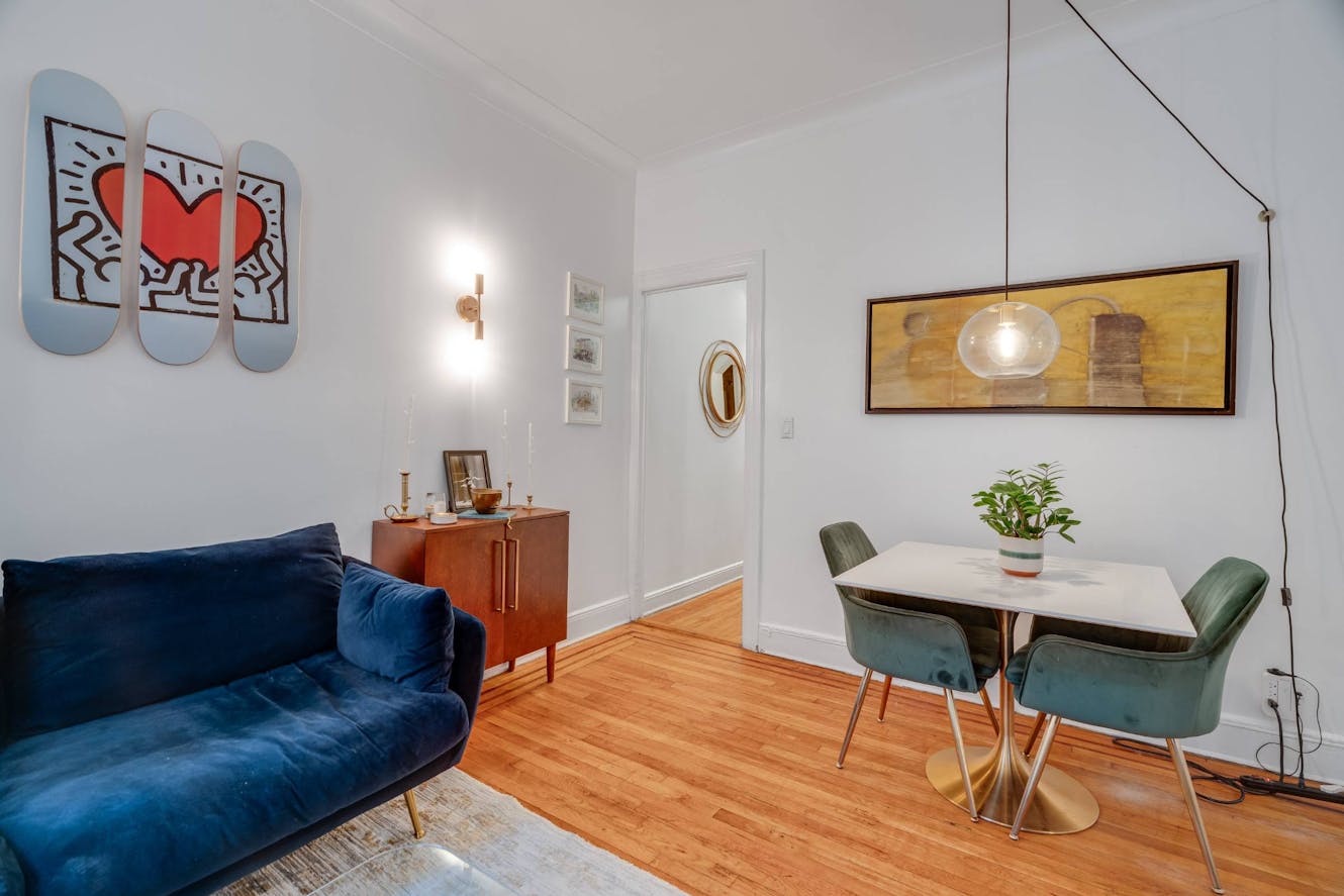 Dining area with white round table, green velvet chairs, arc floor lamp, and blue velvet sofa with wooden credenza and modern artwork
