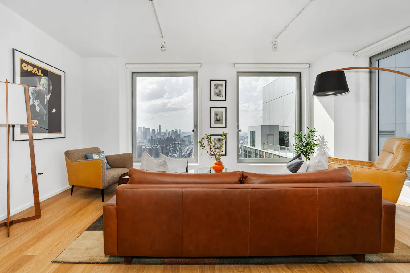 Living room with brown leather sofa, tan accent chairs, large windows with city skyline views, hardwood floors, and modern arc floor lamp