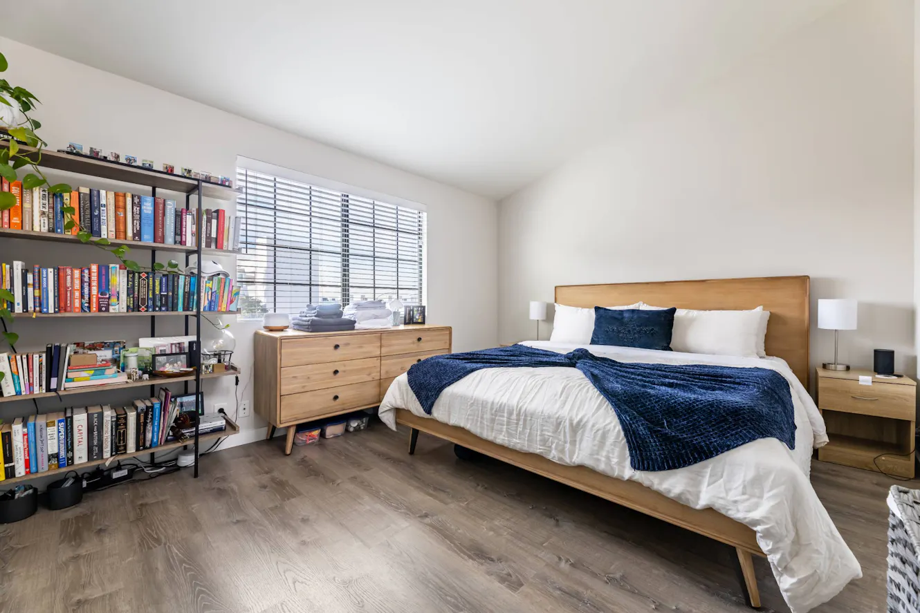 Bedroom with platform bed, white bedding with navy throw, wooden dresser beneath window with blinds, bookshelf with books, and matching nightstands with lamps