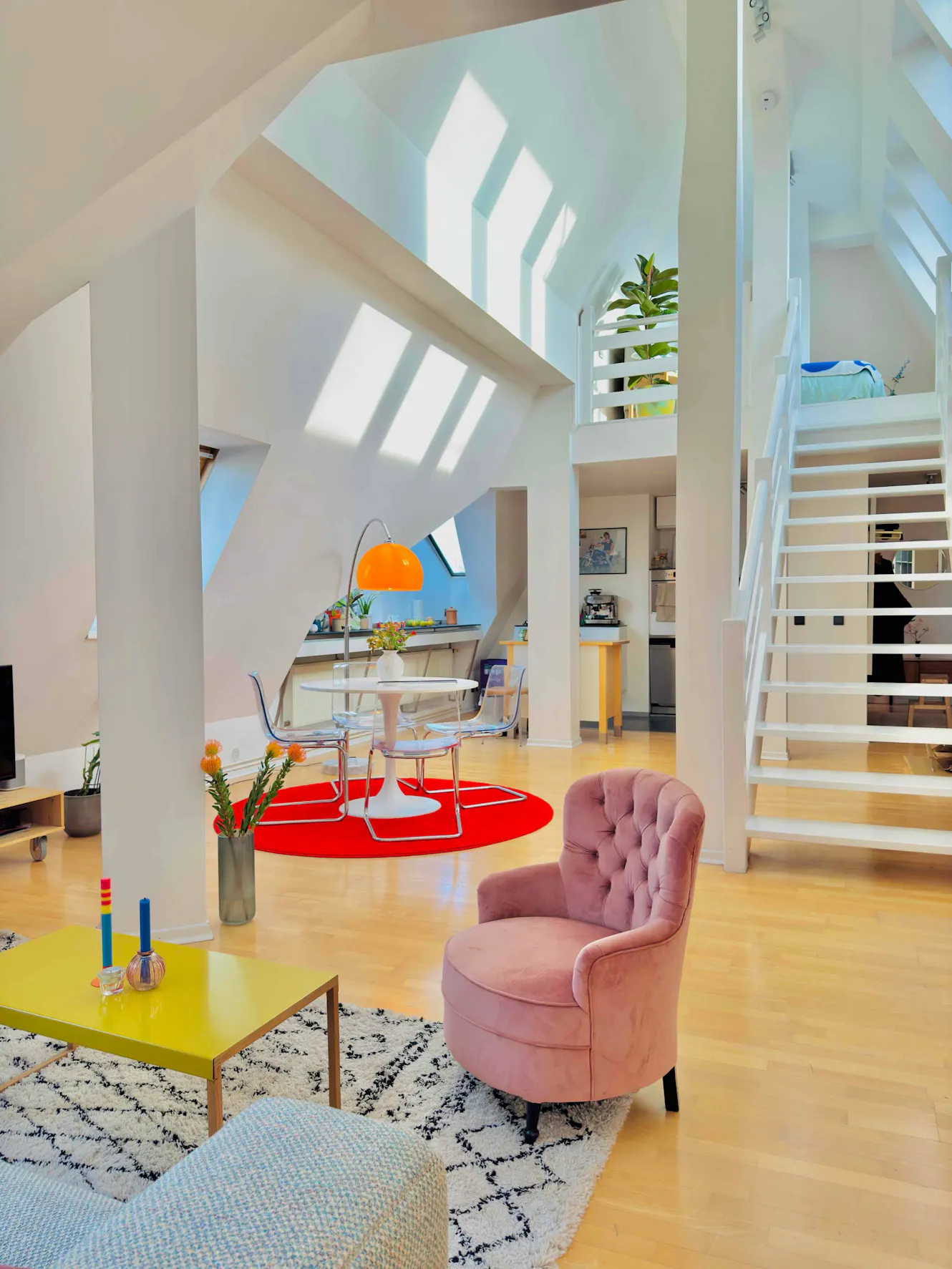 Living room with pink velvet armchair, yellow coffee table, dining area with orange pendant light, white staircase, and skylights with natural light