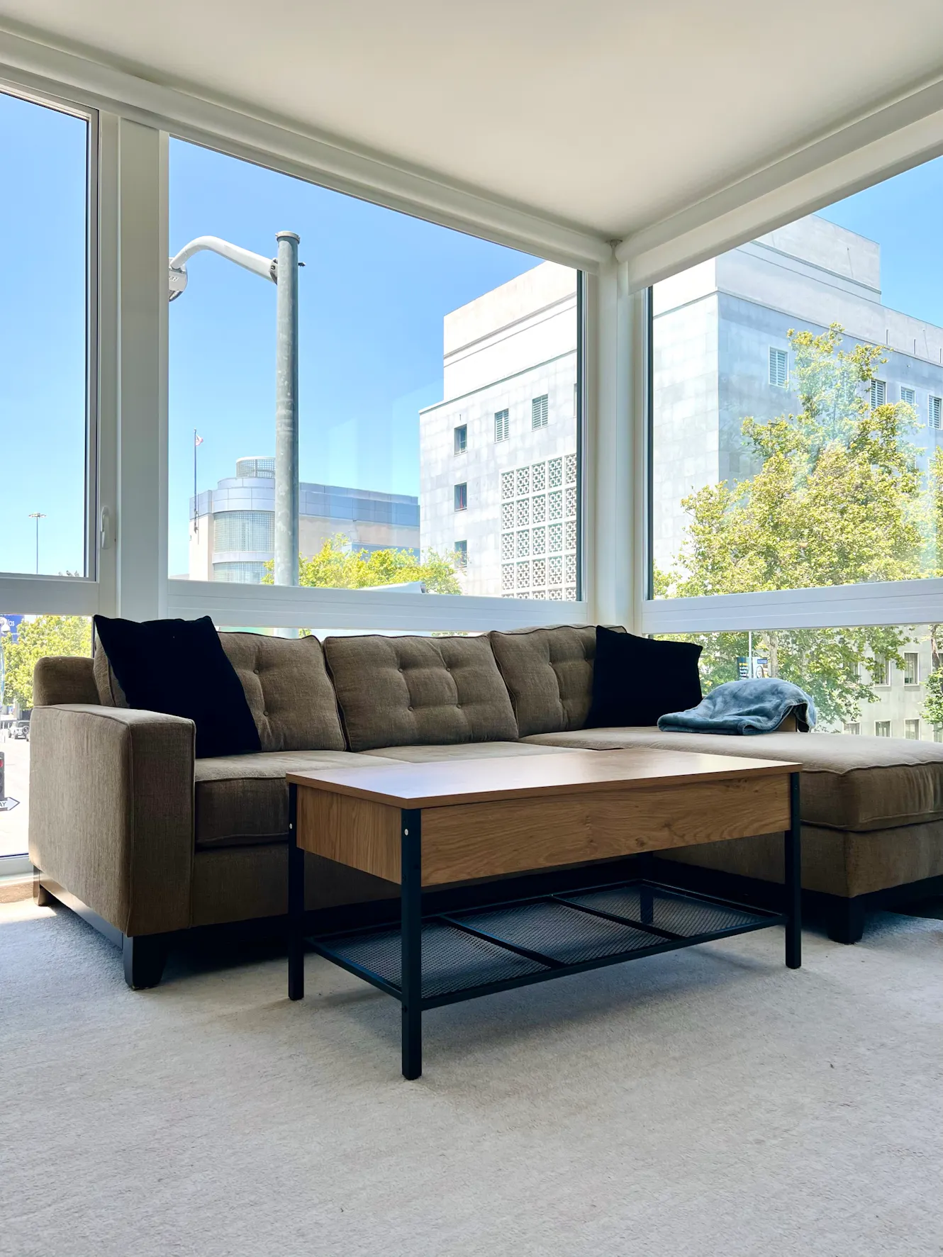 Living room with gray sectional sofa, black accent pillows, wooden coffee table, and floor-to-ceiling windows with city views