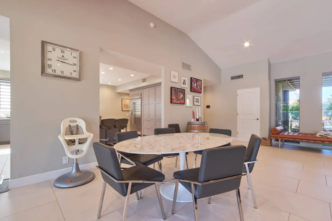 Dining room with round marble table, six gray chairs, high vaulted ceiling, wall clock, and view into kitchen with stainless steel appliances