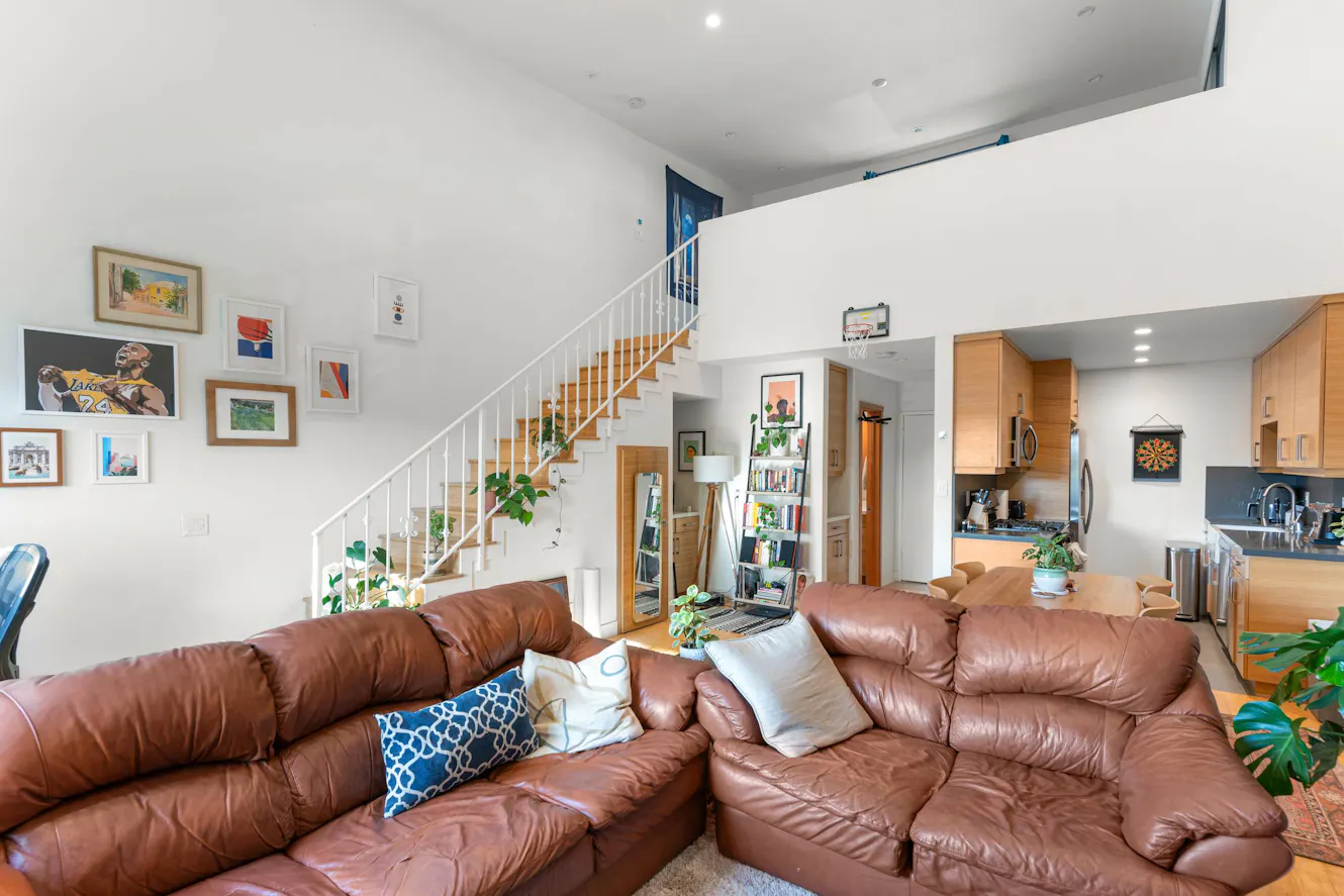 Living room with brown leather sectional sofa, white staircase with wooden steps, gallery wall with framed artwork, and open kitchen with wood cabinets