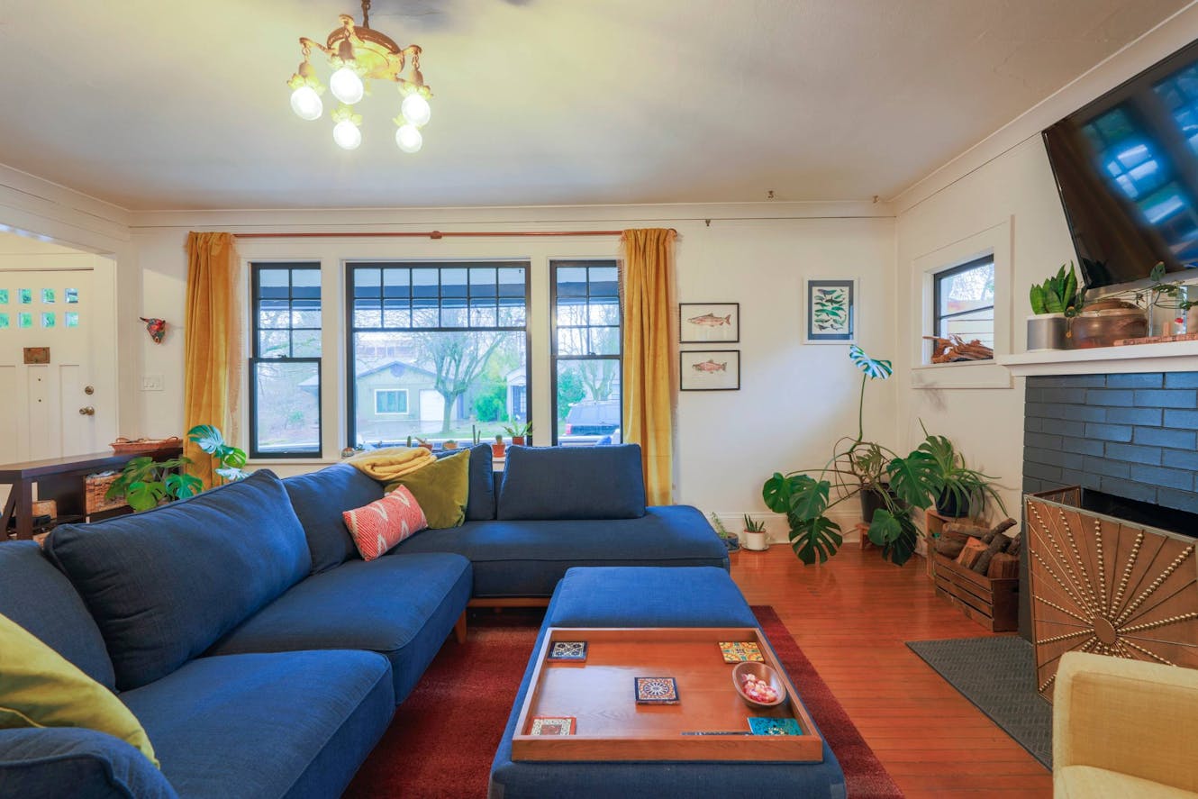 Living room with blue sectional sofa, large windows with natural light, wall-mounted TV above fireplace, hardwood floors, and indoor plants
