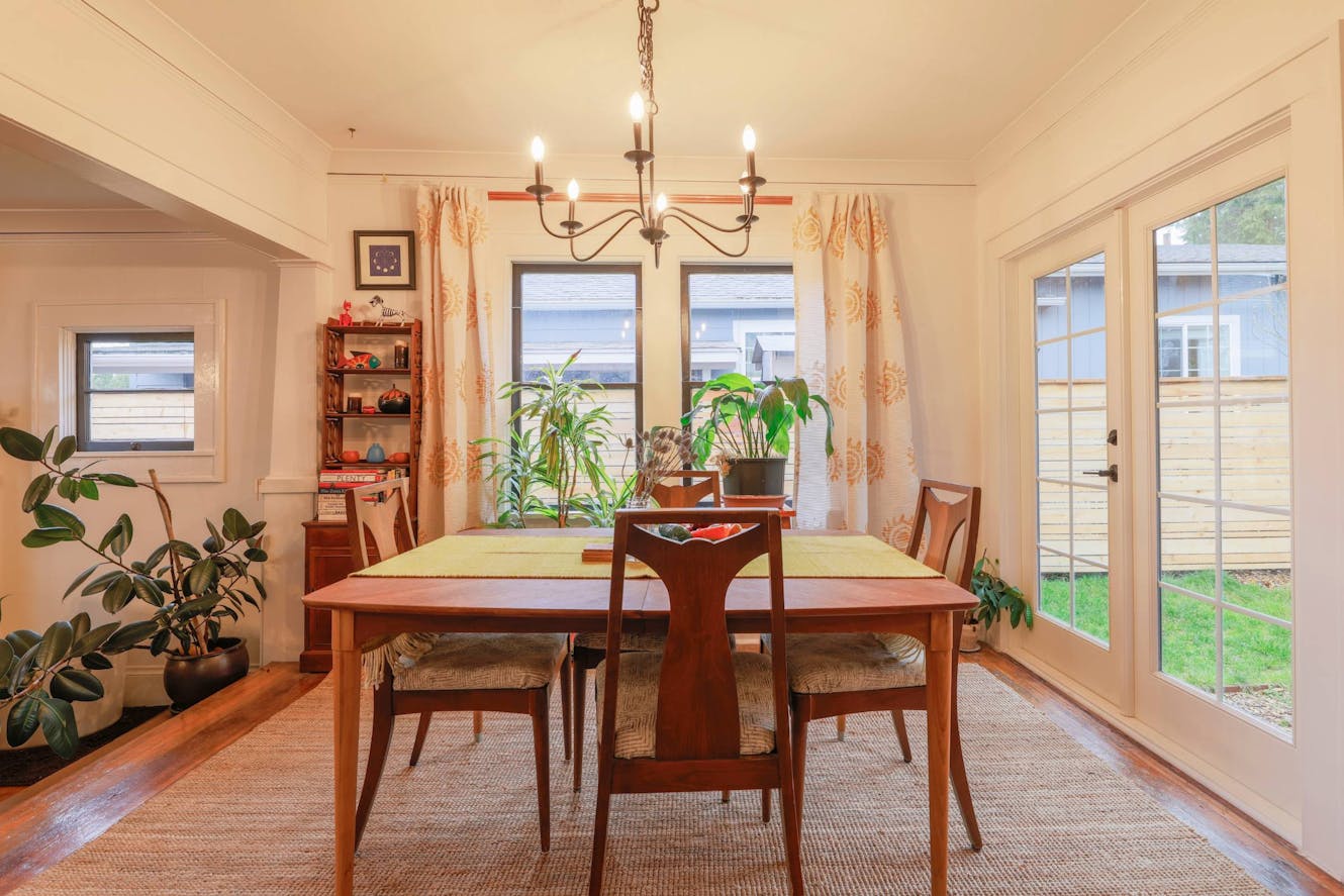 Dining room with wooden table and four chairs, chandelier, patterned curtains, potted plants, and glass door leading to backyard