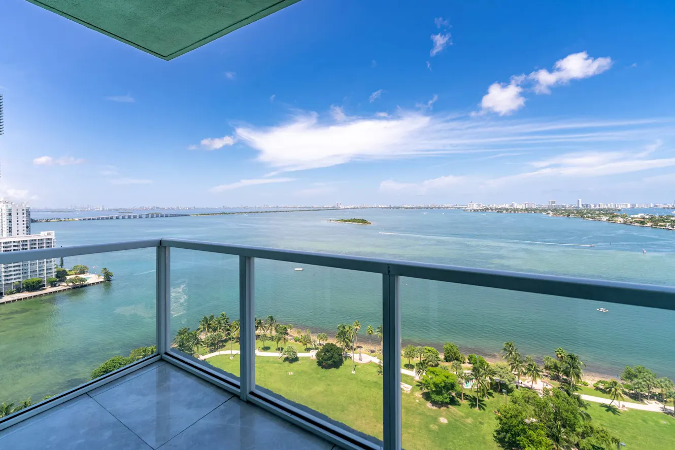 Balcony with glass railing overlooking expansive bay view with bridge, palm trees, green park area, and distant city skyline under blue sky