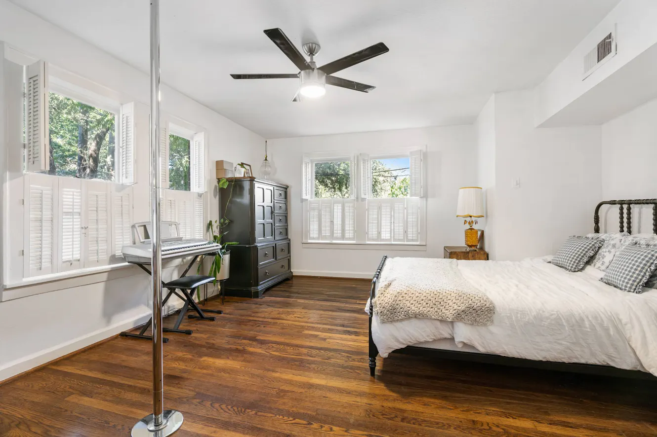 Bedroom with dark wood bed frame, white bedding, dark wood dresser, keyboard workstation, plantation shutters on multiple windows, ceiling fan, and hardwood floors