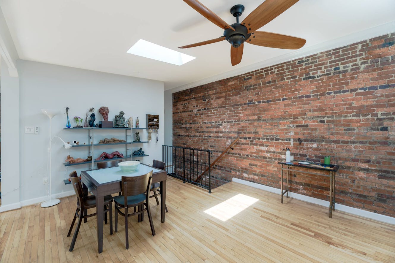 Dining area with exposed brick wall, wooden table with four chairs, ceiling fan, skylight, and staircase railing
