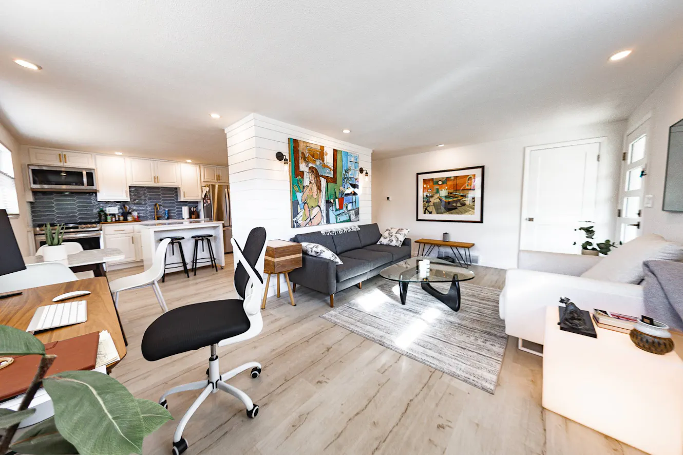 Open-concept living room and kitchen with gray sofa, glass coffee table, white cabinets, stainless steel appliances, light wood flooring, and modern artwork