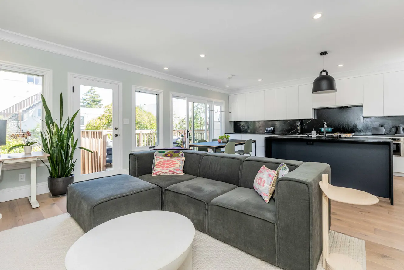 Open-concept living room and kitchen with gray sectional sofa, white coffee table, modern black and white kitchen with island, and glass doors leading to deck