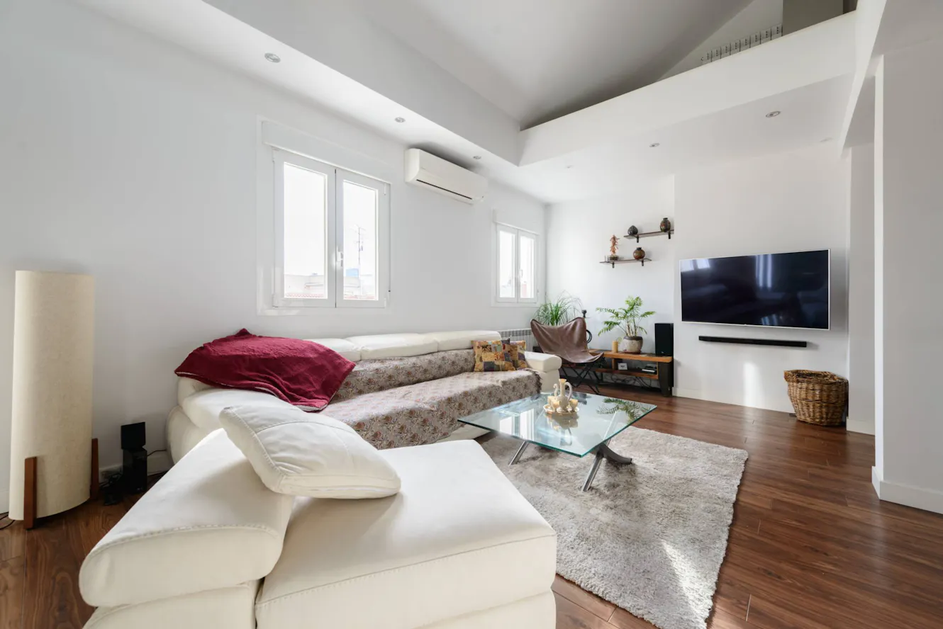 Living room with white sectional sofa, glass coffee table, wall-mounted TV, air conditioning unit, and windows with natural light