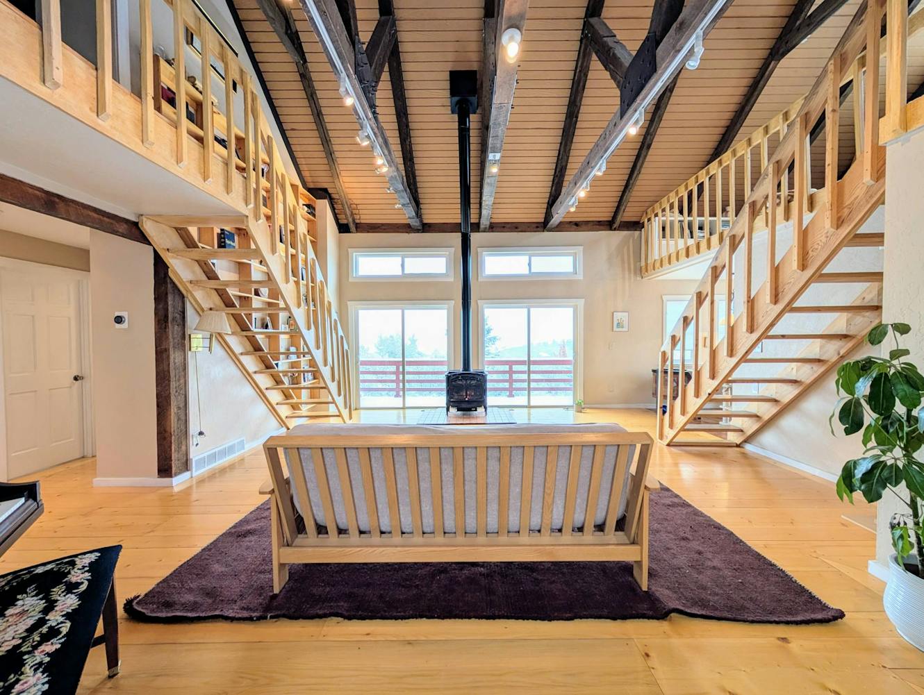 Living room with high vaulted ceiling with exposed beams, wooden staircases to loft areas on both sides, wooden bench on purple rug, large windows, and light wood flooring