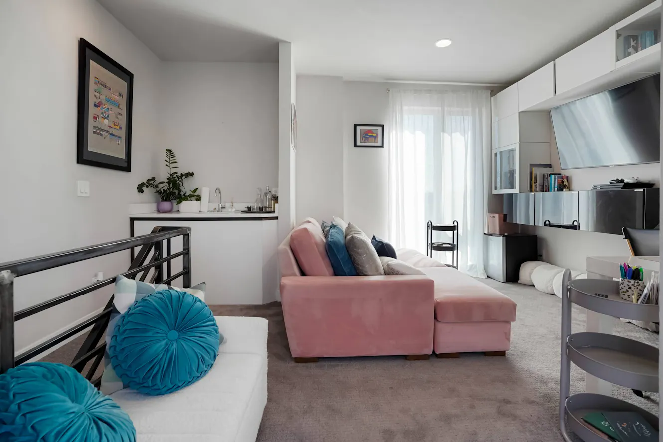 Living room with pink sectional sofa, white dining table with black chairs, white sheer curtains, wall-mounted TV, and workspace area
