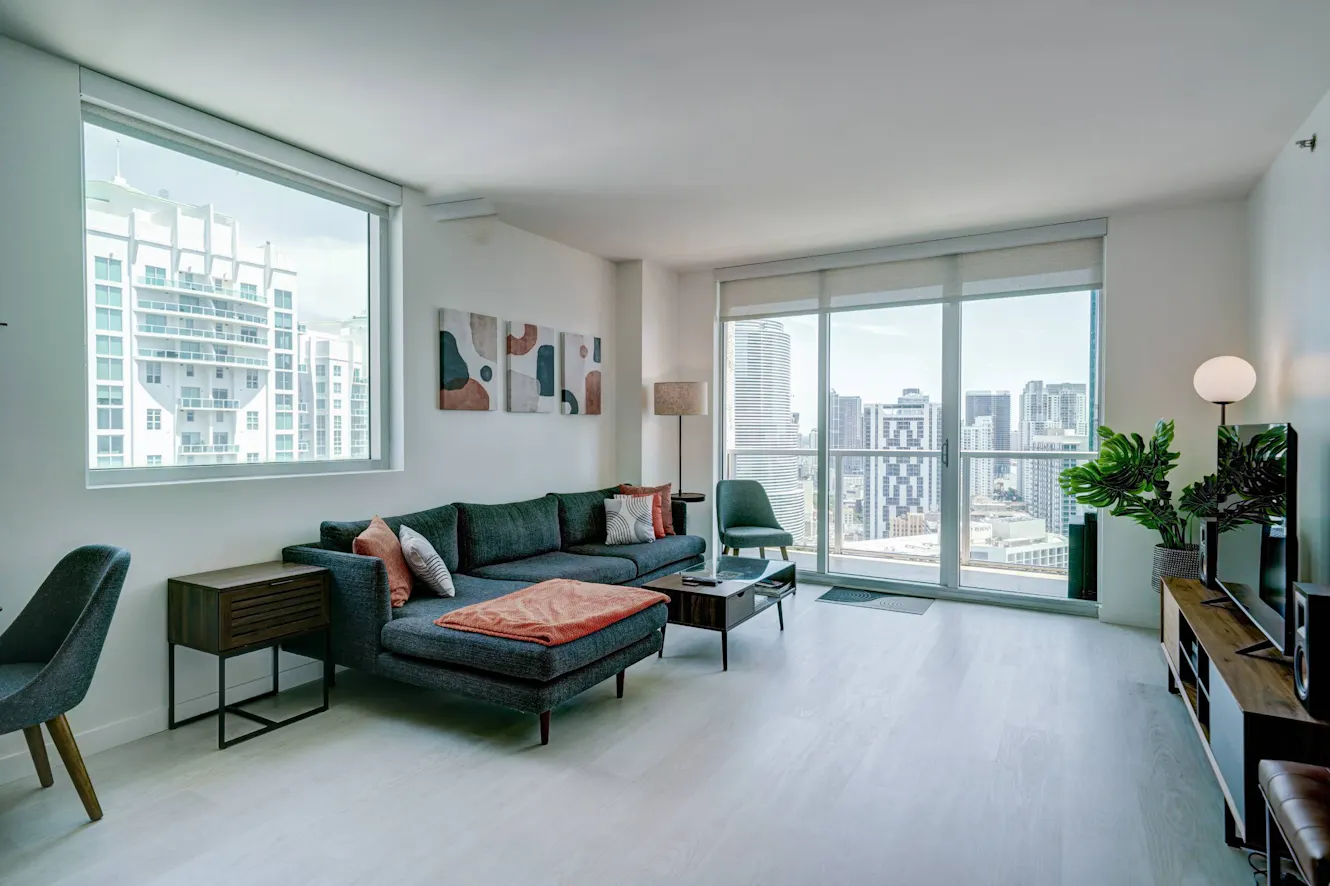 Living room with dark teal sofa and ottoman, floor-to-ceiling windows with city skyline views, modern floor lamp, and indoor plants