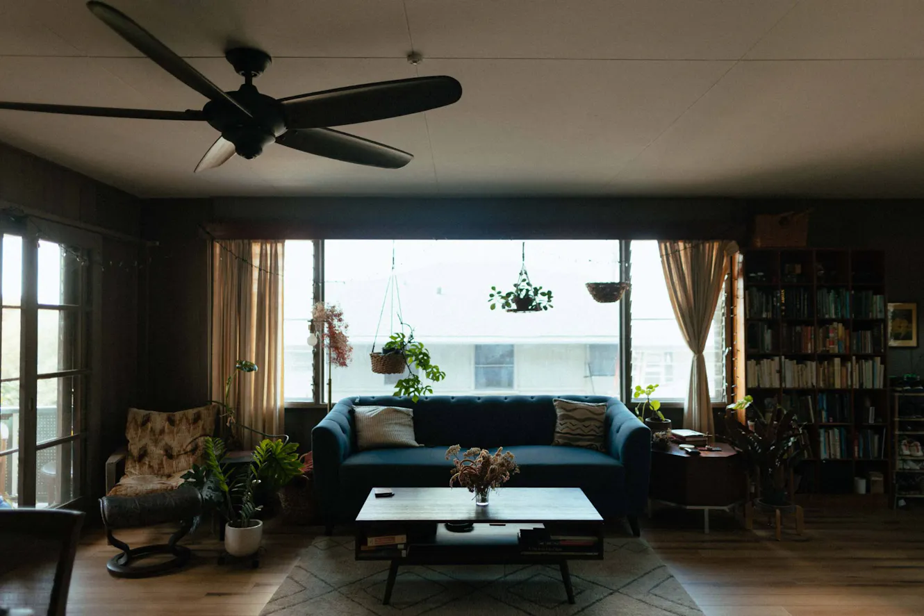 Living room with blue sofa, coffee table with flower vase, ceiling fan, large windows with hanging plants, bookshelf, and armchair