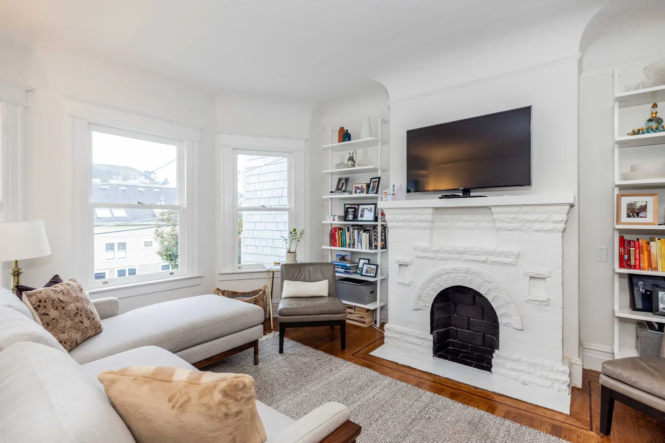 Living room with white painted brick fireplace, wall-mounted TV, gray sofa, two large windows with natural light, built-in shelving, and hardwood floors