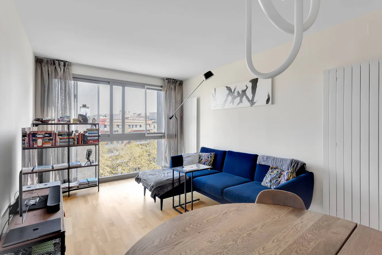 Living room with blue velvet sofa, floor-to-ceiling windows with city views, modern arc floor lamp, bookshelf, and light wood flooring