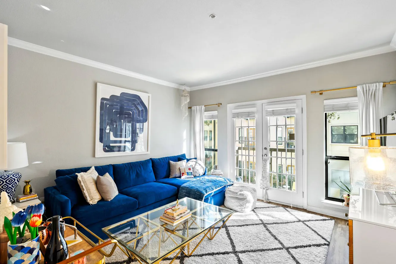 Living room with blue velvet sectional sofa, glass coffee table, white French doors leading to balcony, abstract artwork, and patterned area rug