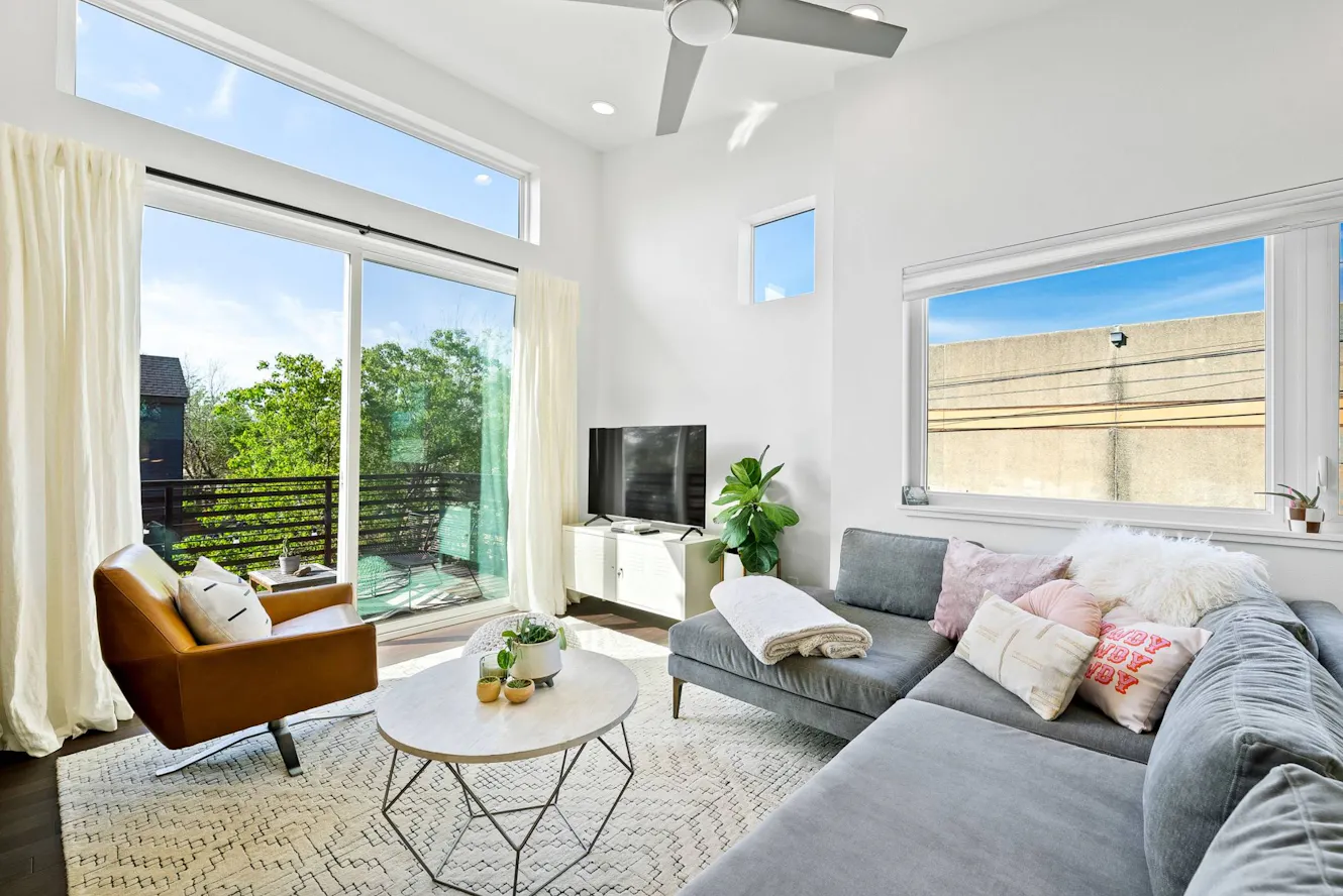Living room with gray sectional sofa, brown leather chair, round coffee table, TV console, ceiling fan, and large windows with natural light and patio access