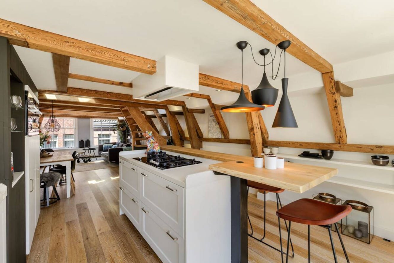 Kitchen with white island featuring gas cooktop, wooden breakfast bar with stools, exposed wooden beams, modern pendant lights, and open view to living area