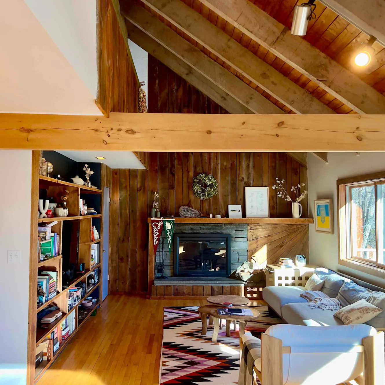 Living room with vaulted wood ceiling, exposed beams, stone fireplace, gray sectional sofa, wooden coffee table, built-in bookshelves, and patterned area rug