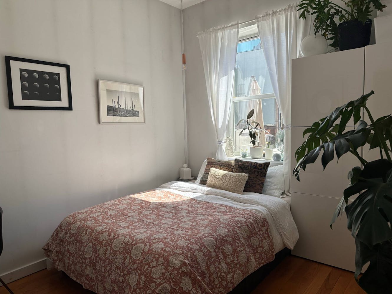 Bedroom with queen bed, white dresser with plants, window with sheer curtains and natural light, framed artwork on walls, and hardwood flooring