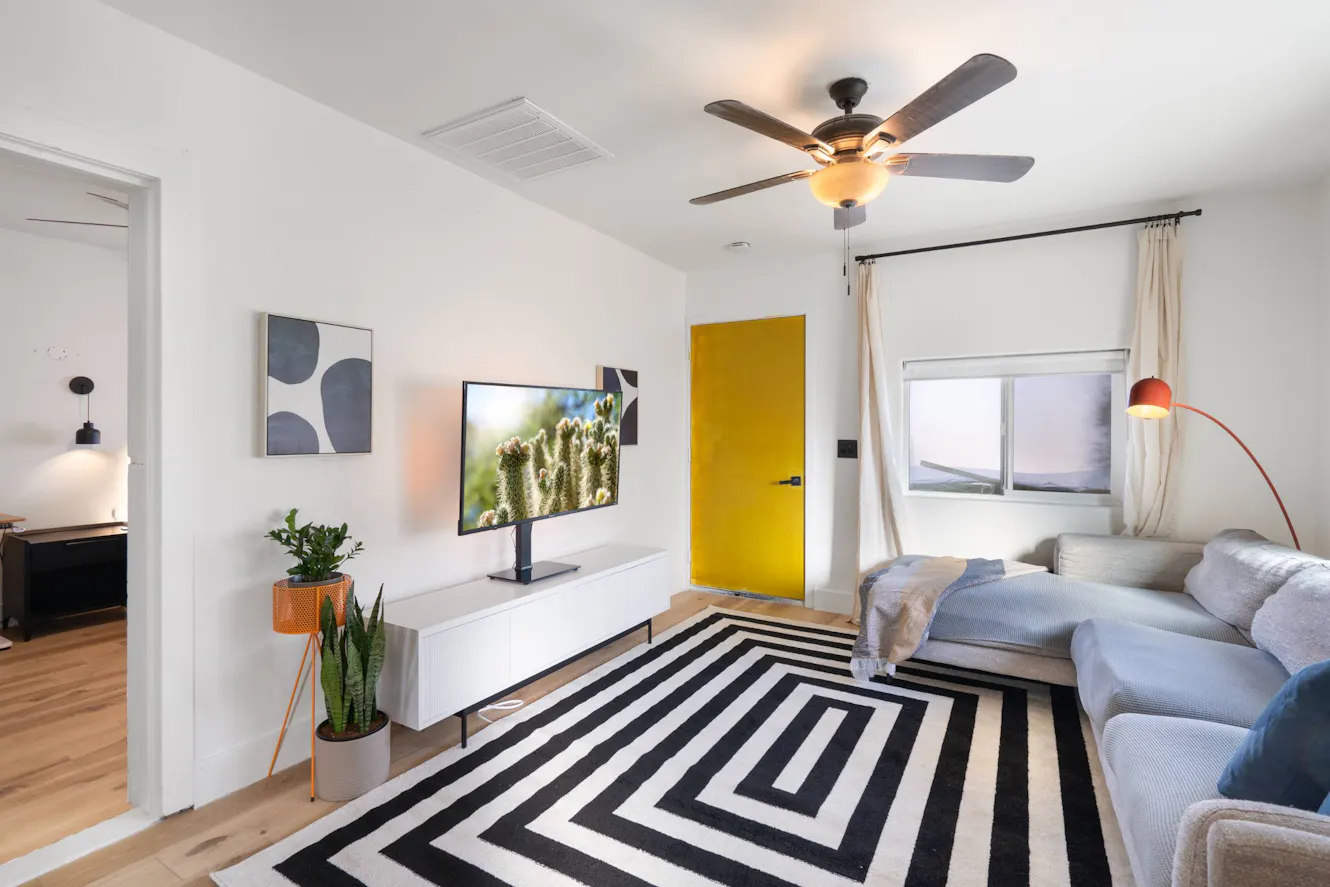 Living room with gray sectional sofa, wall-mounted TV on white media console, ceiling fan with light, geometric black and white rug, and window with curtains