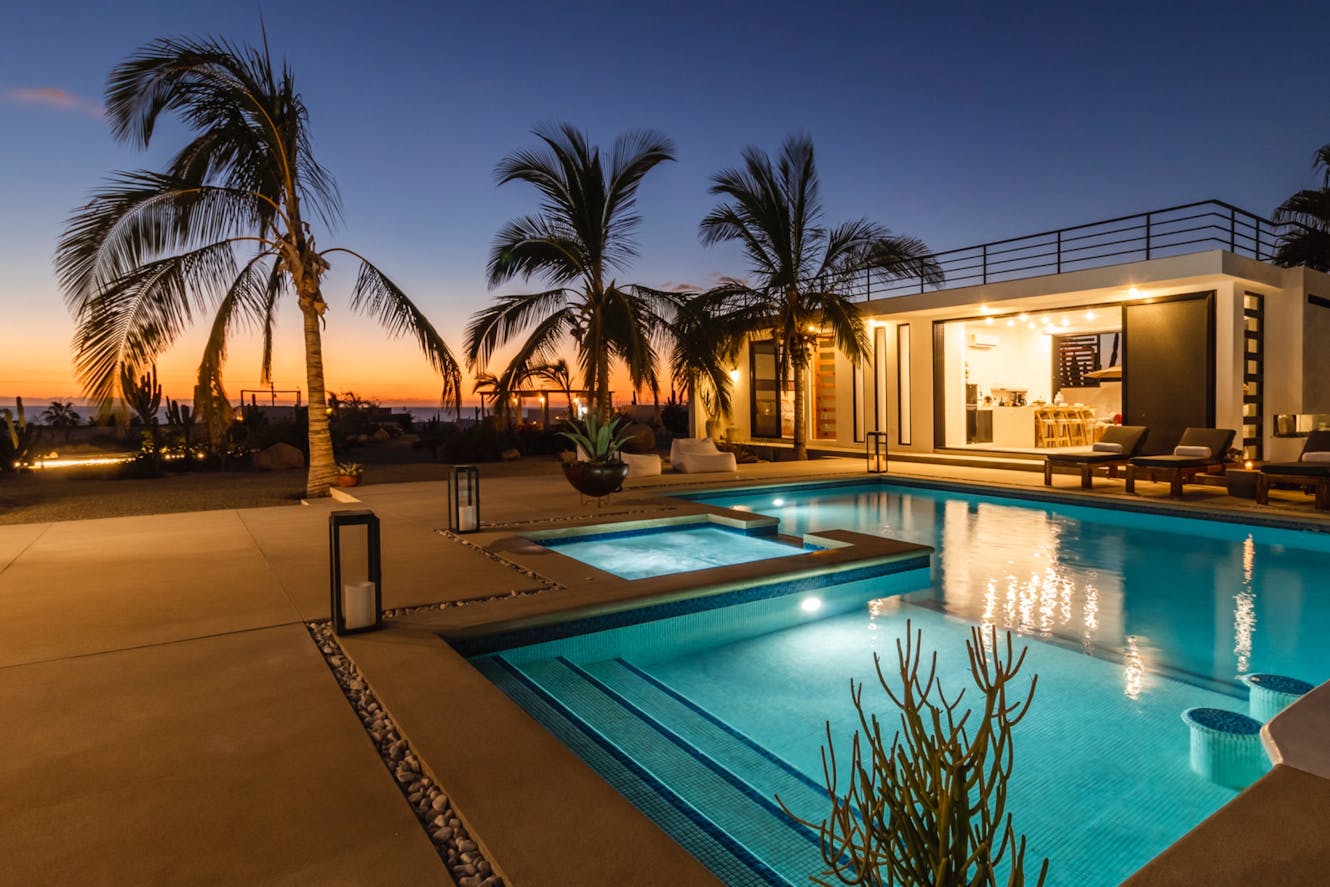 Modern home exterior with illuminated pool, hot tub, palm trees, patio seating, and roof deck at sunset