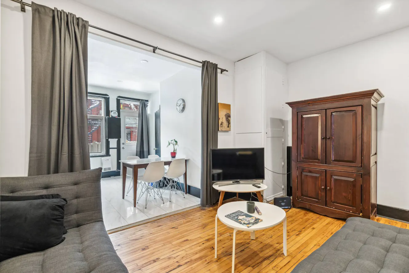 Living room with gray sectional sofa, wooden coffee tables, flat-screen TV, wooden armoire, and view into dining area with white chairs and table