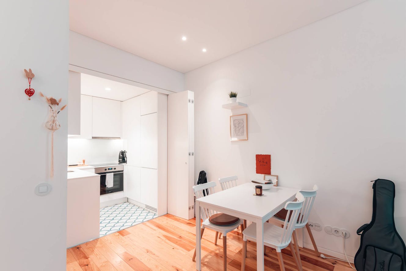 Dining room with white table, four chairs, hardwood floors, and view into modern white kitchen with built-in oven