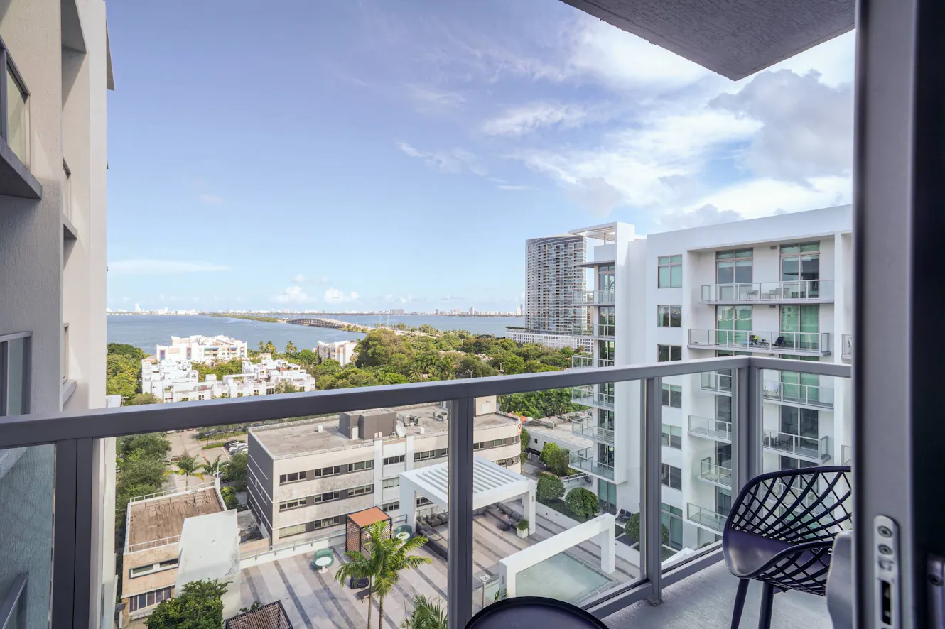 Balcony with black metal railing and chair, overlooking water view with modern buildings, palm trees, and blue sky