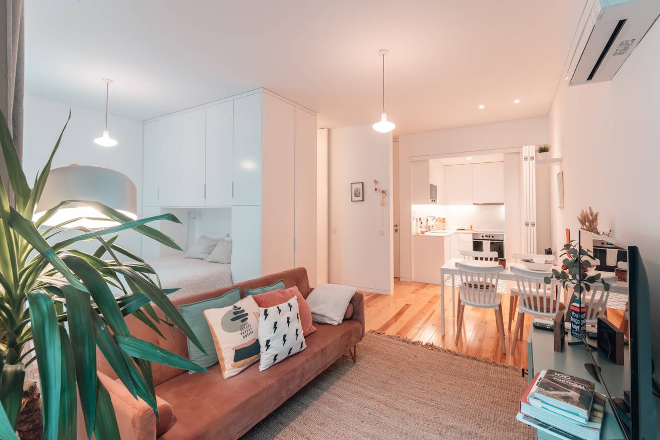 Open-plan living and dining area with rust-colored sofa, white dining table and chairs, hardwood floors, pendant lighting, and view into white kitchen