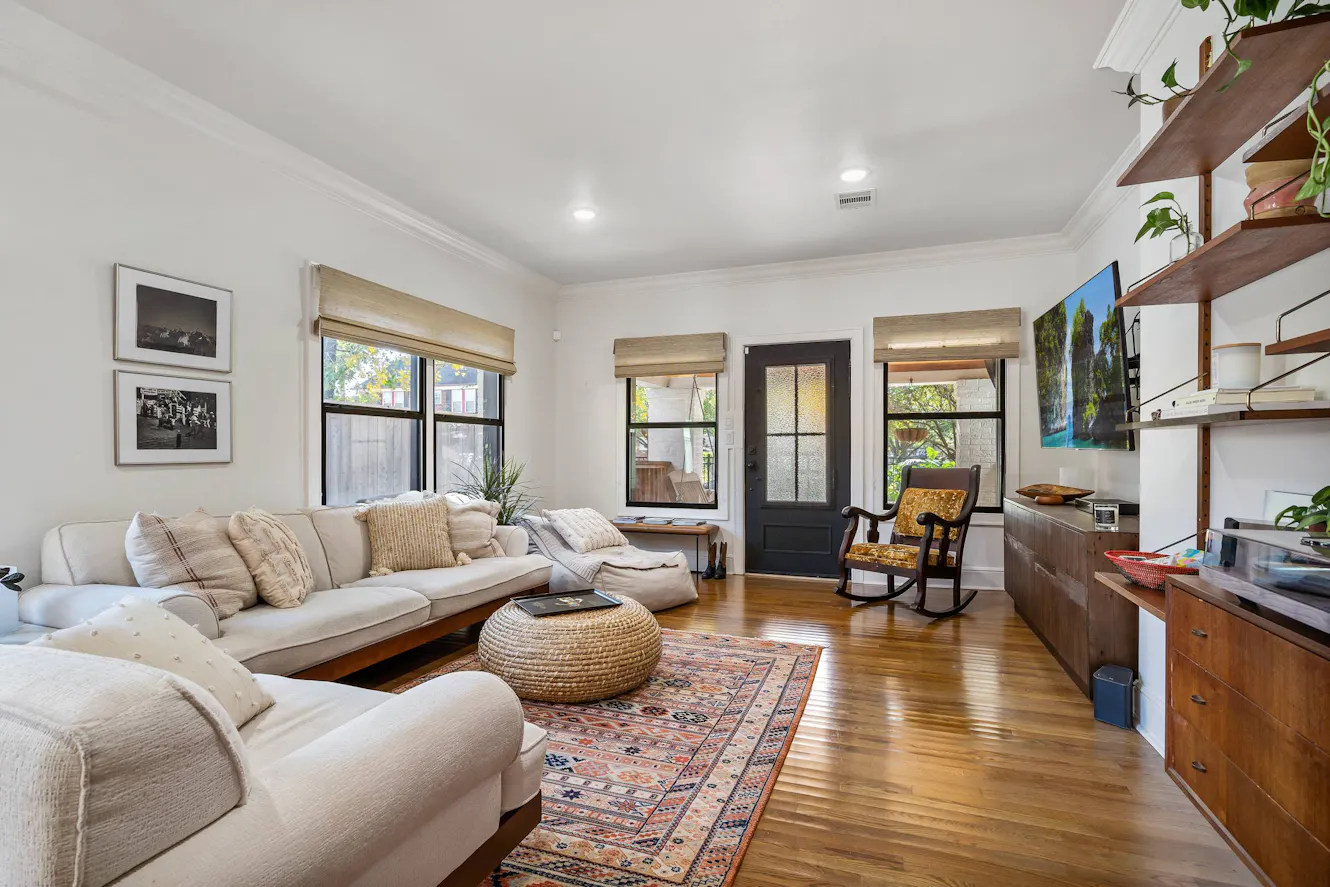 Living room with beige sectional sofa, hardwood floors, multiple windows with natural light, rocking chair, wooden shelving unit, and decorative area rug