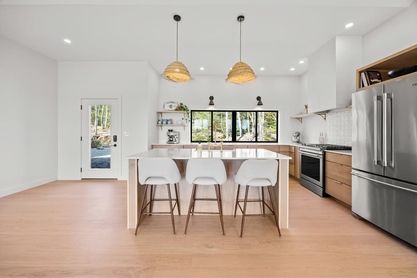 Kitchen with white island, three bar stools, wood cabinets, stainless steel appliances, pendant lights, and large windows with forest view