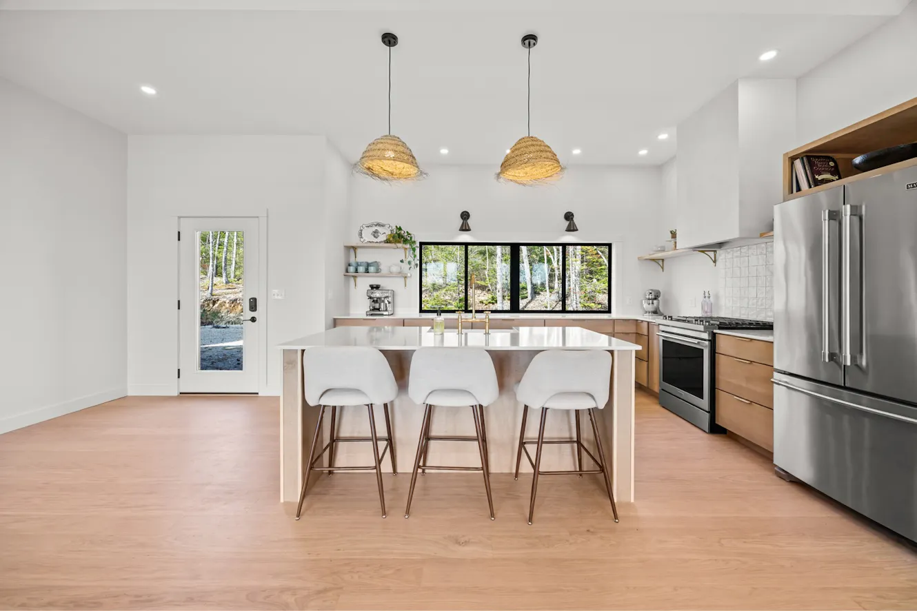 Kitchen with white island, three bar stools, wood cabinets, stainless steel appliances, pendant lights, and large windows with forest view